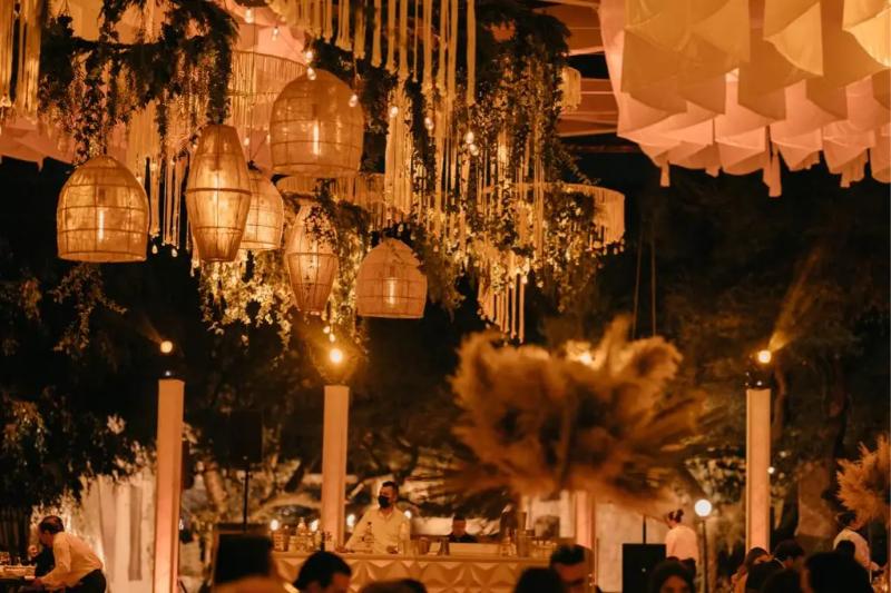 Lámparas de mimbre y telas suspendidas sobre una recepción de noche, con velas altas en las mesas
