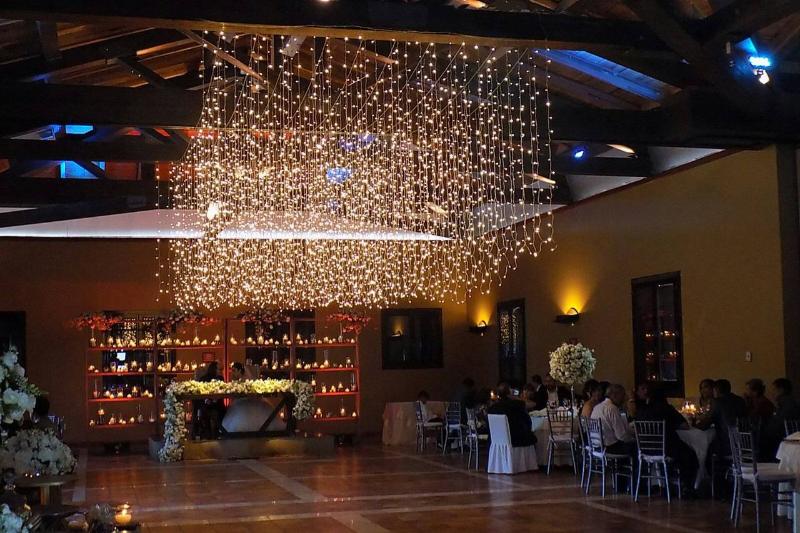 Salón con una cascada de luces colgada del techo, muros iluminados en ámbar y mesas con centros florales
